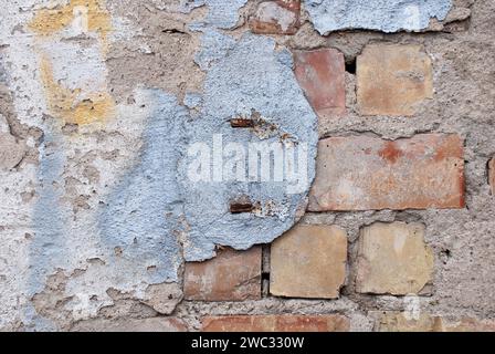 Eine verwitterte Mauer mit Überresten von Gips und freiliegenden Ziegeln, alte Mauer aus roten Ziegeln mit blauer, weißer und gelber Farbe auf Gipsresten, Symbol für Stockfoto