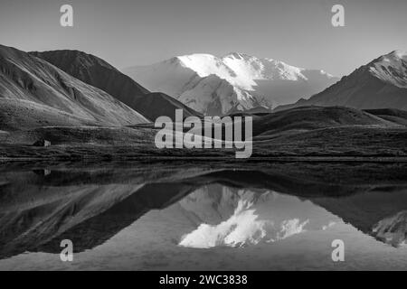 Weiß vergletschter und schneebedeckter Berggipfel Lenin Peak bei Sonnenuntergang, Berge spiegeln sich in einem See zwischen goldenen Hügeln, Schwarz-weiß-Foto, Trans Stockfoto