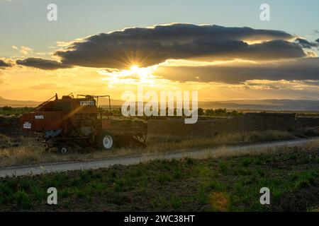 Ein Oldtimer-Mähdrescher steht ungenutzt auf einem trockenen Feld unter einem dramatischen Sonnenuntergang mit Bergen in der Ferne Stockfoto