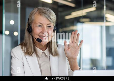Professionelle, reife Frau mit einem Headset, das freundlich winkt und für Kundensupport und positive Interaktion am Arbeitsplatz steht. Stockfoto