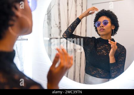 Afrikanische Frau mit Sonnenbrille kämmt ihre Haare und macht sich bereit für die Nachtparty vor dem Spiegel Stockfoto