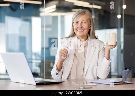 Porträt einer lächelnden, grauhaarigen Geschäftsfrau, die im Büro am Tisch sitzt, mit einer Tasse und einer Serviette und in die Kamera lächelt. Stockfoto