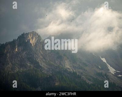 Atmosphärischer Blick auf die italienischen Alpen über Courmayeur mit schwachem Sonnenschein, der durch die niedrigen Wolken durchdrungen ist und über den schneebedeckten Gipfeln und Felsen kreisen Stockfoto