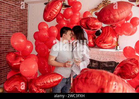 Fröhlicher Moment zwischen jungen Paaren, die mit Toast den Valentinstag in der Nähe von roten Ballons und weißem Kamin feiern. Frau lacht, hält Glas, kurz vor C Stockfoto