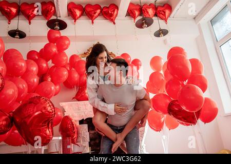 Verspielter, romantischer Moment zwischen verrücktem Paar in der Nähe von roten Ballons in einem hellen Raum mit weißer Inneneinrichtung. Frau sitzt auf dem Rücken des Mannes. Ein paar, das herumspielt Stockfoto