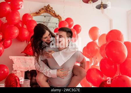 Verspielter, romantischer Moment zwischen verrücktem Paar in der Nähe von roten Ballons in einem hellen Raum mit weißer Inneneinrichtung. Frau sitzt auf dem Rücken des Mannes. Ein paar, das herumspielt Stockfoto