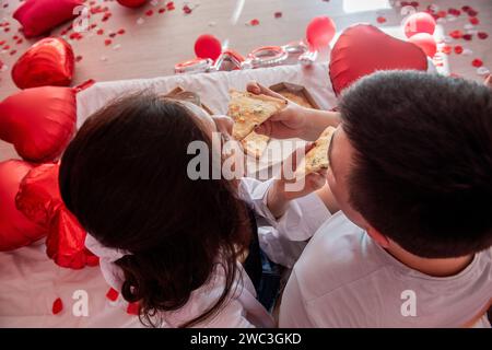 Blick von oben auf junge Paare, die sich gegenseitig füttern, Pizza aus der Take-away-Box genießen, mit Gläsern Champagner und grünen Trauben. Mann und Frau teilen sich festlich, Stockfoto