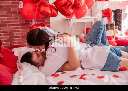 Verliebtes Paar umarmt sich eng auf weißem Bett zwischen roten Ballons, Kissen, Herzen, Rosenblüten. Mann und Frau sehen sich an, verbringen den Valentinstag zu Hause Stockfoto