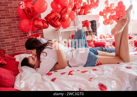 Verliebtes Paar umarmt sich eng auf weißem Bett zwischen roten Ballons, Kissen, Herzen, Rosenblüten. Mann und Frau sehen sich an, verbringen den Valentinstag zu Hause Stockfoto