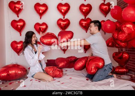 Verspielter Moment zwischen Paaren, die sich in Kissenballons auf dem Bett kämpfen, mit herzförmigen roten Ballons im Hintergrund. Die Frau macht fröhlich rumtäuscht Stockfoto