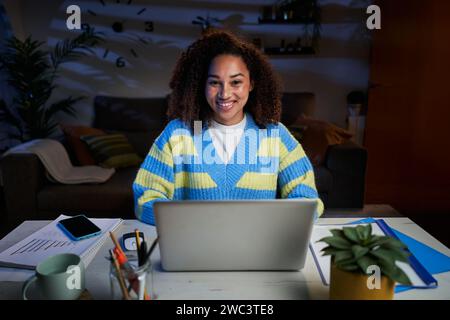 Porträt attraktive junge Afro-Frau, die in die Kamera lächelnd vor dem Laptop zu Hause sitzt. Stockfoto