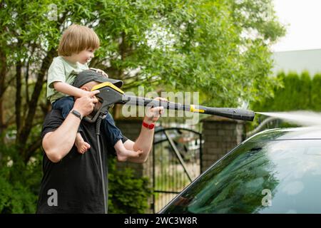 Vater und sein kleiner Sohn benutzen eine Wasserpistole, um ein Auto zu waschen. Männlicher Fahrer, der ein Auto mit kontaktlosem Hochdruckwasserstrahl wäscht. Reinigen eines Fahrzeugs in Stockfoto