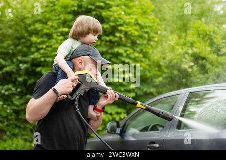 Vater und sein kleiner Sohn benutzen eine Wasserpistole, um ein Auto zu waschen. Männlicher Fahrer, der ein Auto mit kontaktlosem Hochdruckwasserstrahl wäscht. Reinigen eines Fahrzeugs in Stockfoto