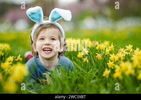 Niedlicher Kleinkind mit Hasenohren, der Spaß zwischen den Reihen wunderschöner gelber Narzissen hat, die am Frühlingstag blühen. Ostern im Freien. Ei h Stockfoto