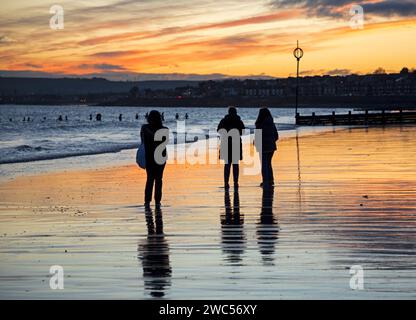 Portobello, Edinburgh, Schottland, Großbritannien. 14. Januar 2024. Zuschauer am Ufer des Firth of Forth bei Sonnenaufgang beobachten die Kaltwasserschwimmer, die ein Bad im Morgengrauen am Meer nehmen. Die Temperatur ist kalt, 2 Grad Celsius. Quelle: Archwhite/Alamy Live News. Stockfoto