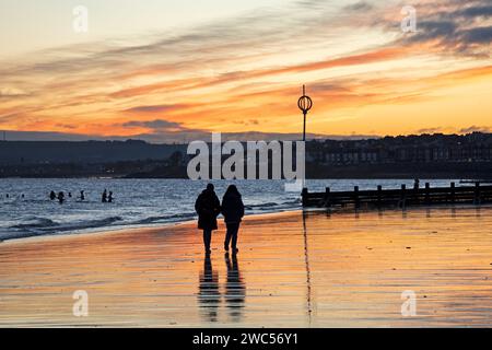 Portobello, Edinburgh, Schottland, Großbritannien. 14. Januar 2024. Zuschauer am Ufer des Firth of Forth bei Sonnenaufgang beobachten die Kaltwasserschwimmer, die ein Bad im Morgengrauen am Meer nehmen. Die Temperatur ist kalt, 2 Grad Celsius. Quelle: Archwhite/Alamy Live News. Stockfoto