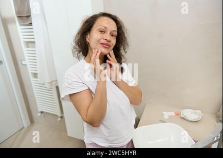 Attraktive Frau mit lockigen Haaren mittleren Alters im weißen Pyjama, lächelnd und blickend in die Kamera, während sie feuchtigkeitsspendenden Reinigungsschaum auf ihr Gesicht aufträgt, s Stockfoto