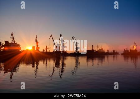 Silhouetten bei Sonnenuntergang, die Kräne im geschäftigen Hafen transportieren. Kommerzielle Frachtdocks während der goldenen Stunde, Sonnenstrahlen auf dem Wasser. Welthandel, Güterverkehr Stockfoto