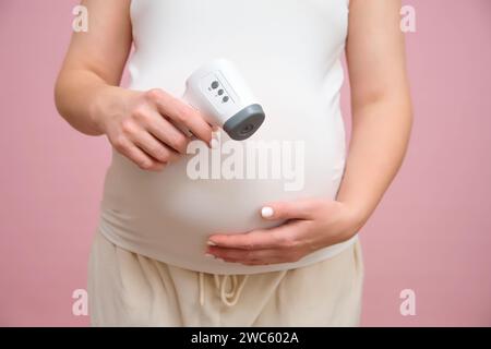 Schwangere Frau, die ein berührungsloses elektronisches Thermometer in der Hand hält, rosafarbener Hintergrund Stockfoto
