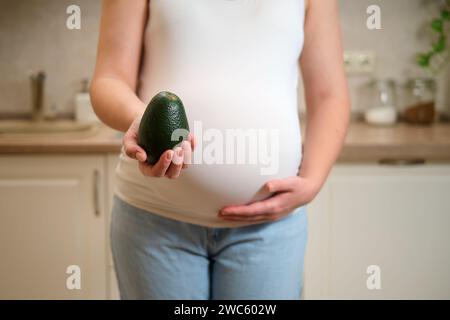 Eine schwangere Frau hält ein Avocadogemüse in den Händen, während sie in einer Küche steht. Schwangerschaft und richtige Ernährung während des Wartens auf ein Baby Stockfoto