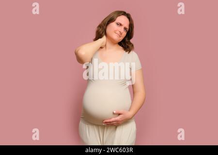 Eine schwangere Frau hält sich an einem schmerzenden Hals auf einem rosa Hintergrund des Studios fest. Schwangerschaft bei einer Frau mit Bauch Stockfoto