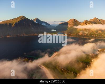 Aus der Vogelperspektive der Lofoten, mit Wolken darunter, Sonnenuntergang und Orangentönen. Blauer Himmel oben. Fjord, Seen und Berggipfel. Stockfoto