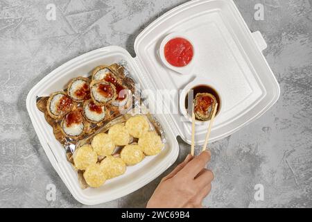 Köstliche Sushi-Rollen in Einweg-Plastikboxen. Die Hand nimmt eine gebackene Rolle mit Essstäbchen. Service-Konzept für die Bereitstellung von Lebensmitteln, flach stapelbar Stockfoto