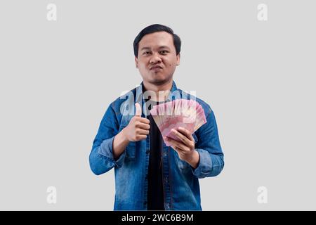 Junger asiatischer Mann mit arrogantem Gesichtsausdruck, während er Papiergeld hält und einen Daumen nach oben gibt Stockfoto