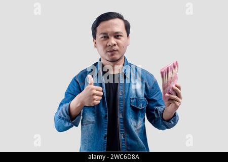 Junger asiatischer Mann mit stolzem Gesichtsausdruck, während er Papiergeld hält und einen Daumen nach oben gibt Stockfoto