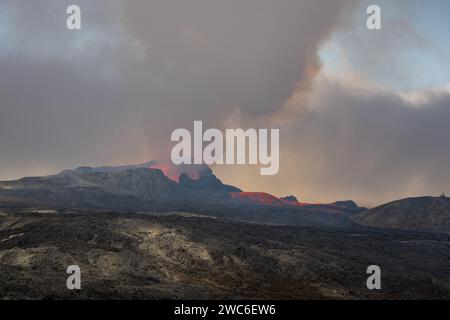 Blick über ein gehärtetes schwarzgraues Lavafeld. Durch schimmernde heiße Luft brechen Lava und Rauch aus einem Vulkan in der Ferne aus. Stockfoto