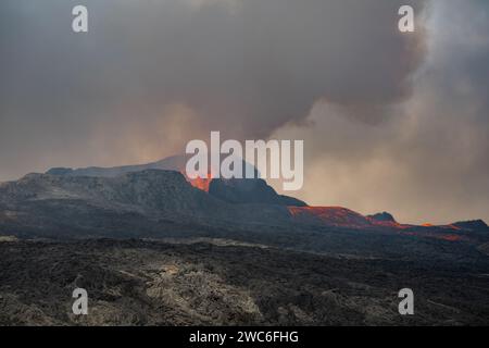 Blick über ein gehärtetes schwarzgraues Lavafeld. Durch schimmernde heiße Luft brechen Lava und Rauch aus einem Vulkan in der Ferne aus. Stockfoto