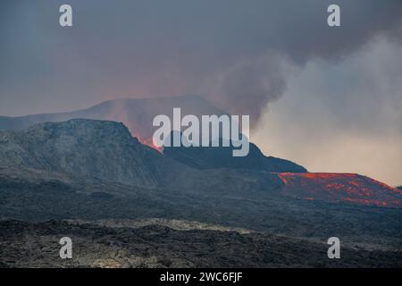 Blick über ein gehärtetes schwarzgraues Lavafeld. Durch schimmernde heiße Luft brechen Lava und Rauch aus einem Vulkan in der Ferne aus. Stockfoto