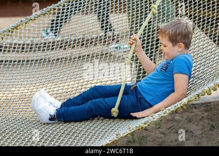 Porträt eines kleinen Jungen in Kletterausrüstung in einem Seilpark, der ein Seil mit einem Knoten hält. Das Kind spielt im Seilpark. Hindernislauf für Kinder. Stockfoto