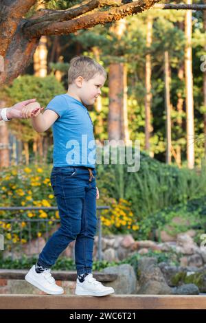 Ein konzentrierter Junge läuft mit einem Holzbalken darauf. Sonniger Sommergarten im Hintergrund. Stockfoto