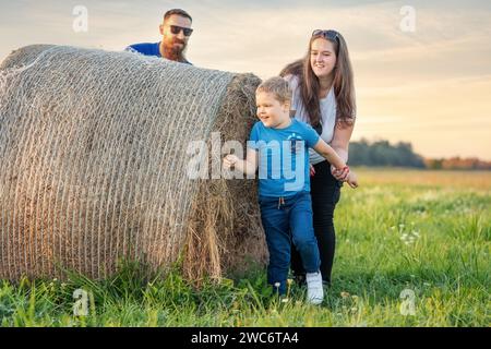 Sommerferienabend lustige Familienspiele auf der Wiese um den Heuballen. Mutter und Sohn fangen Vater und er versteckt sich hinter einer Rolle Stockfoto