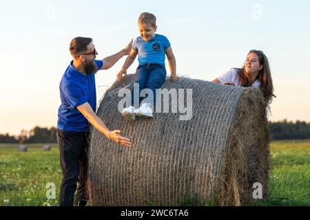 Ein fröhlicher kleiner Junge rutscht einen runden Heuballen hinunter, sein Vater versucht, ihn zu fangen. Eine fröhliche, schöne junge Familie ist mit ihrem Kleinkind verlobt Stockfoto