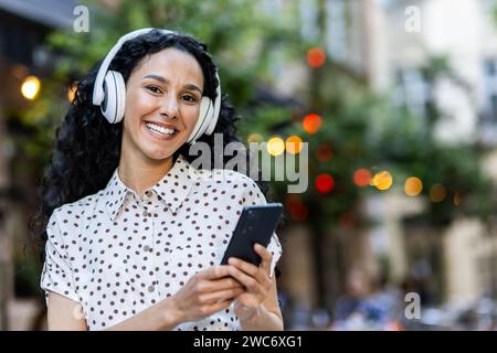 Fröhliche Frau mit Kopfhörern, die ihr Smartphone draußen nutzt. Fröhliches, vernetztes, urbanes Lebenskonzept mit Bokeh-Lichtern im Hintergrund. Stockfoto