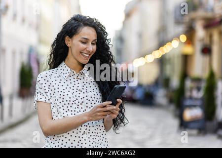 Lächelnde Frau, die ihr Smartphone auf einer belebten Straße nutzt und Konnektivität, Freude und urbanen Lebensstil in einem unscharfen Hintergrund ausdrückt. Stockfoto