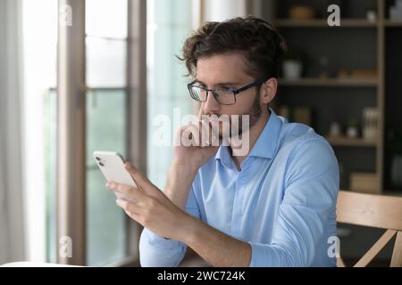 Ein ernster Mann sitzt am Schreibtisch und liest die Nachricht auf dem Handy Stockfoto