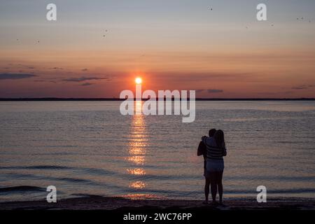 Zwei Individuen stehen an einem Sandstrand und betrachten den faszinierenden Anblick der Sonne, die unter dem Horizont untergeht Stockfoto