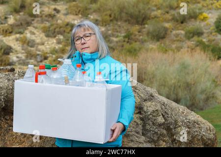 Ältere weißhaarige Frau, die eine Pappschachtel voller leerer Plastikflaschen hält, die auf dem Feld zum Recycling gesammelt wurden, Konzept der Ökologie Stockfoto