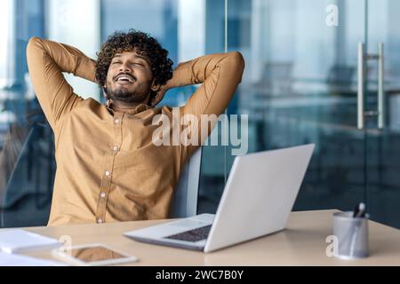 Zufriedener junger Mann mit lockigen Haaren, der sich in seinem Bürostuhl zurücklehnt, die Augen geschlossen und die Hände hinter dem Kopf, um einen friedlichen Moment zu genießen. Stockfoto