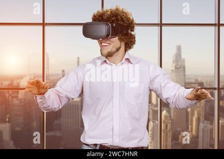 Porträt eines glücklichen jungen, fröhlich aufgeregten Mannes mit lockigem Haar, der vr-Headset trägt. Innenfenster mit Blick auf die Stadt am Abend im Hintergrund. Stockfoto