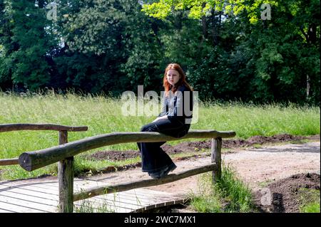 Ein rothaariges Teenager-Mädchen sitzt traurig auf dem hölzernen Geländer einer Brücke in grüner Natur an einem sonnigen Sommertag Stockfoto