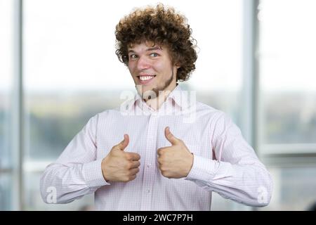 Ein glücklicher, lächelnder junger Mann mit lockigen Haaren zeigt beide Daumen hoch. Innenfenster im Hintergrund. Stockfoto