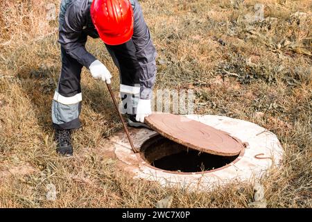 Der Klempner in einem Helm hebt die Mannlochabdeckung eines Wasserbrunnens an. Inspektion und Wartung von Sanitär- und Wasserzählern. Stockfoto