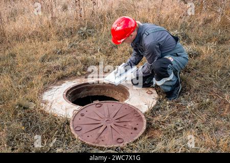 Klempner in Overalls und ein Helm in der Nähe eines Wasserbrunnens schreibt die durchgeführten Messungen und die Messwerte des Wasserzählers auf. Stockfoto