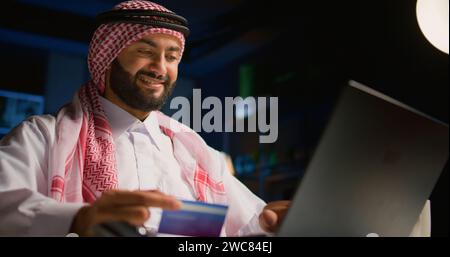 Fröhlicher arabischer Mann, der im Internet einkaufen und Kreditkarteninformationen am Computer eingibt. Lächelnde Person aus dem Nahen Osten, die auf der Website eine Zahlungsmethode hinzufügt, bezahlt für Online-Bestellung von Lebensmitteln Stockfoto