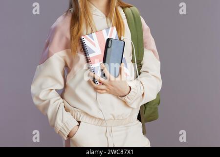 Nahaufnahme eines Teenager-Mädchens in beigefarbenem Trainingsanzug mit Rucksack, englischer Flagge und Kopfhörern mit Smartphone-Anwendungen auf grauem Hintergrund. Stockfoto