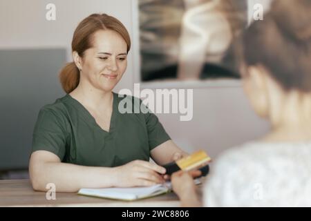 Gesundheitszeit. Glückliche Massage-Therapeutin in Massagekabine mit Klientin, die Kreditkartenzahlungen über das Terminal an der Rezeption akzeptiert. Stockfoto